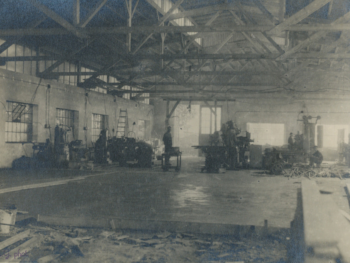 Die Werkhalle in Strullendorf ist noch im Rohbau. Der Boden ist teilweise verlegt und die Decke ist noch nicht ausgebaut. Im Hintergrund sieht man Arbeiter, die am Ausbau beteiligt sind, im Jahr 1940.