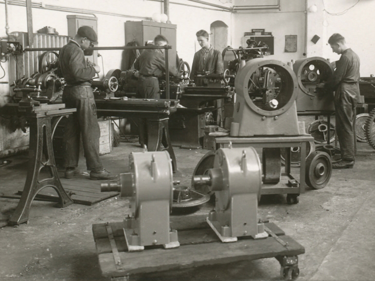 Mehrere Arbeiter stehen an den Werkzeugen und fertigen Zahngetriebe im Jahr 1930 an. Die Arbeiter schauen nach unten und sind in ihre Arbeit vertieft. Die Getriebe stehen rechts auf Rollplatten. KACHELMANN GETRIEBE GmbH