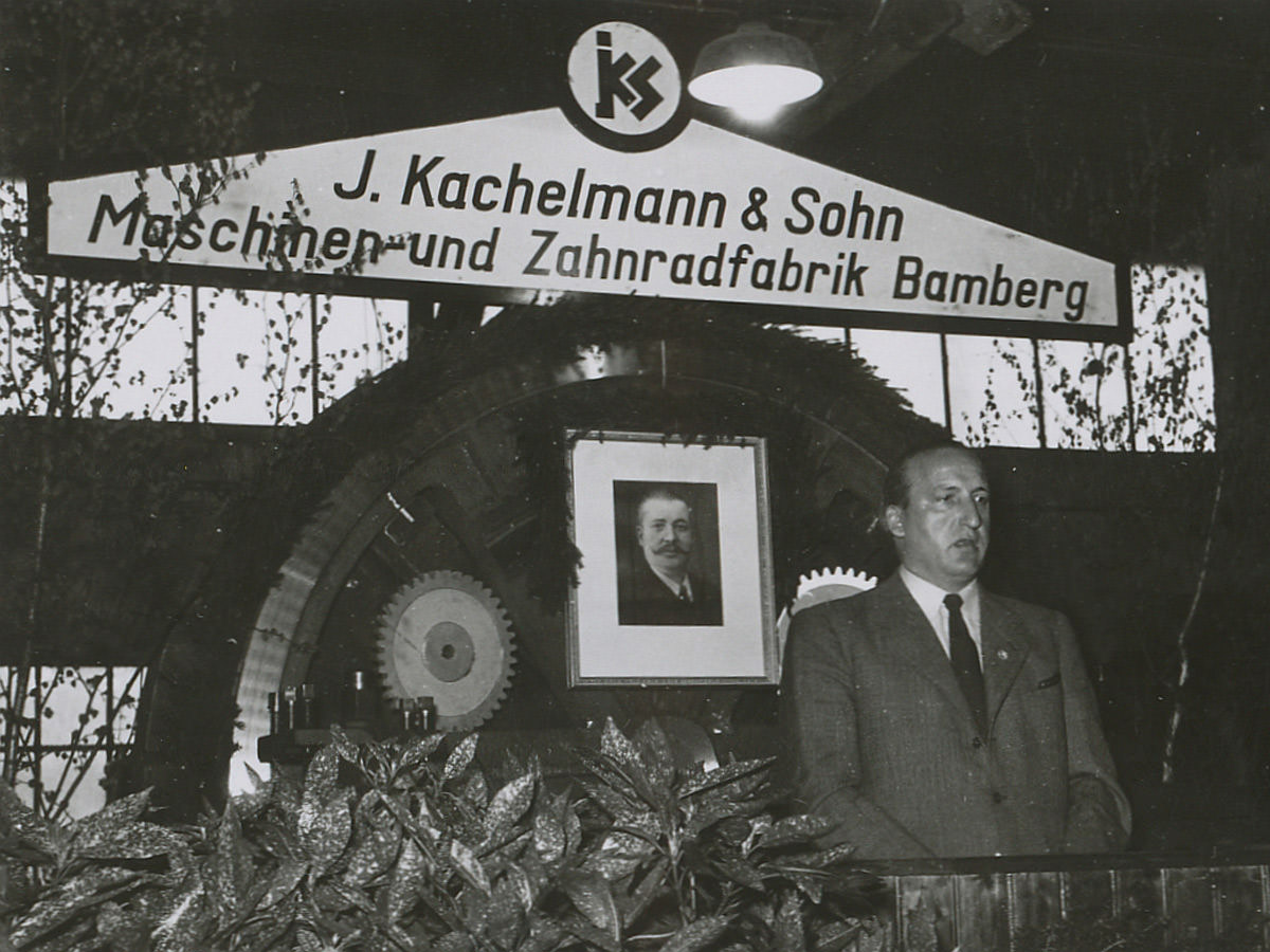 Der Geschäftsführer von KACHELMANN GETRIEBE GmbH steht am Pult und redet. Im Hintergrund ist ein Getriebe zu sehen. Das Bild ist schwarz-weiß und aus dem Jahr 1954.