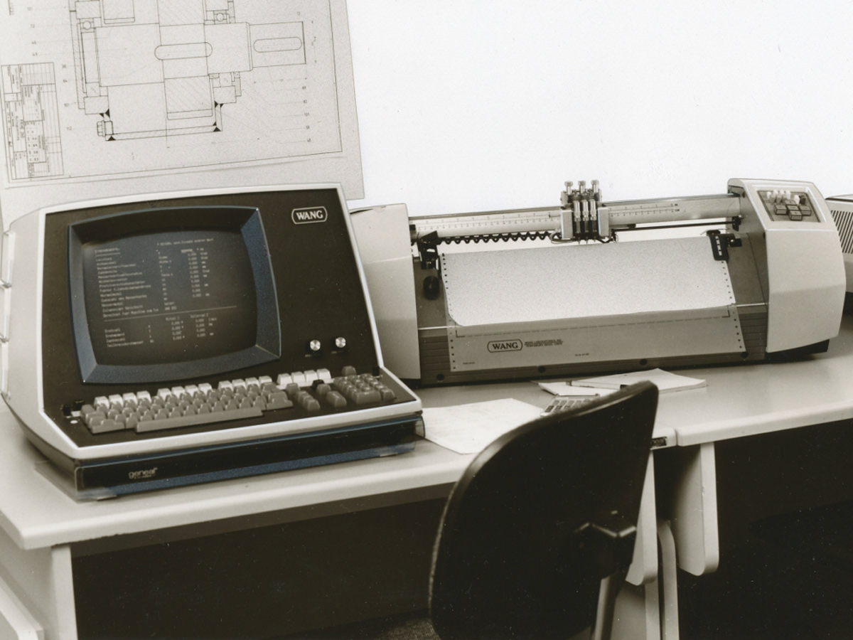 Das Bild zeigt einen modernen Schreibtisch aus dem Jahr 1988. Darauf befinden sich Arbeitsgeräte, wie ein Computer aus dem Jahr 1988. Hinter dem Computer befindet sich eine technische Zeichnung. KACHELMANN GETRIEBE GmbH
