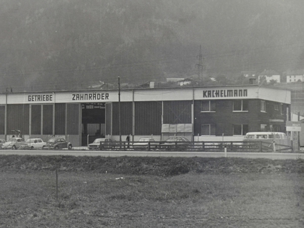 Das Bild zeigt die Montagehalle von KACHELMANN AUSTRIA mit Schlosserwerkstatt im österreichischen Rietz im Jahr 1974. Die Autos parken vor der Werkhalle, davor befindet sich eine Straße und eine Wiese mit Zaun. KACHELMANN GETRIEBE GmbH