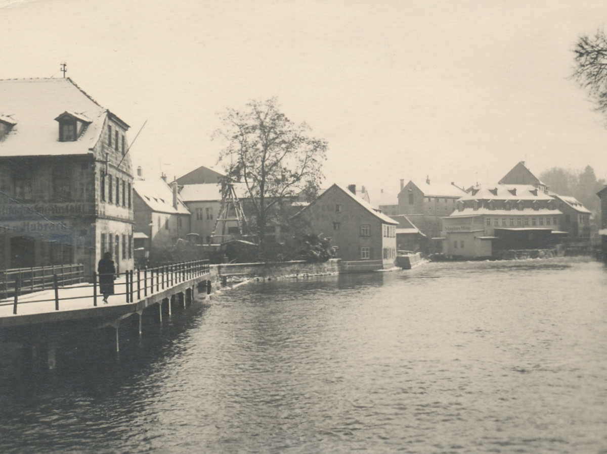 Auf dem historischen Bild sieht man die obere Mühle aus dem Jahr 1941. Im unteren rechten Bereich ist der Fluss in Bamberg zu erkennen und links an der Seite ist ein Gehweg. KACHELMANN GETRIEBE GmbH