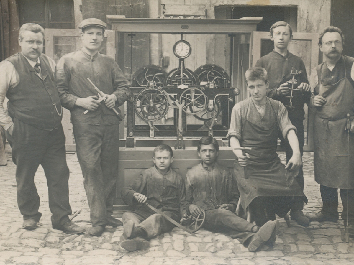 Das schwarz-weiß Bild zeigt eine Turmuhr, die von den KACHELMANN Mitarbeitern im Jahr 1910 repariert wurde. Die Arbeiter halten Werkzeuge in den Händen und sind an die Turmuhr gelehnt. KACHELMANN GETRIEBE GmbH