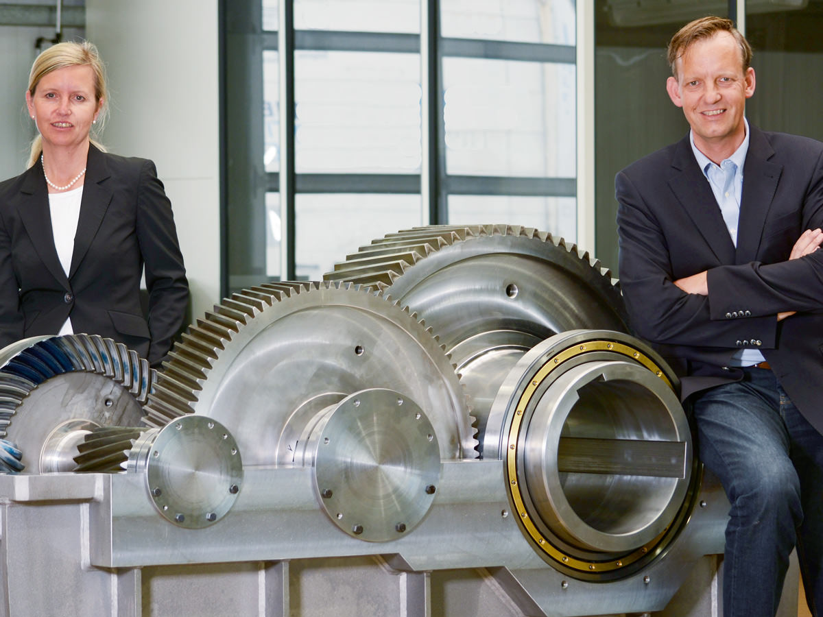 Die beiden Geschäftsführer der Firma KACHELMANN GETRIEBE GmbH, Alexander Kachelmann v. Colberg und Andrea von Wartenberg lehnen an einem Getriebe. Im Hintergrund ist die Werkshalle der Firma zu erkennen. KACHELMANN GETRIEBE GmbH